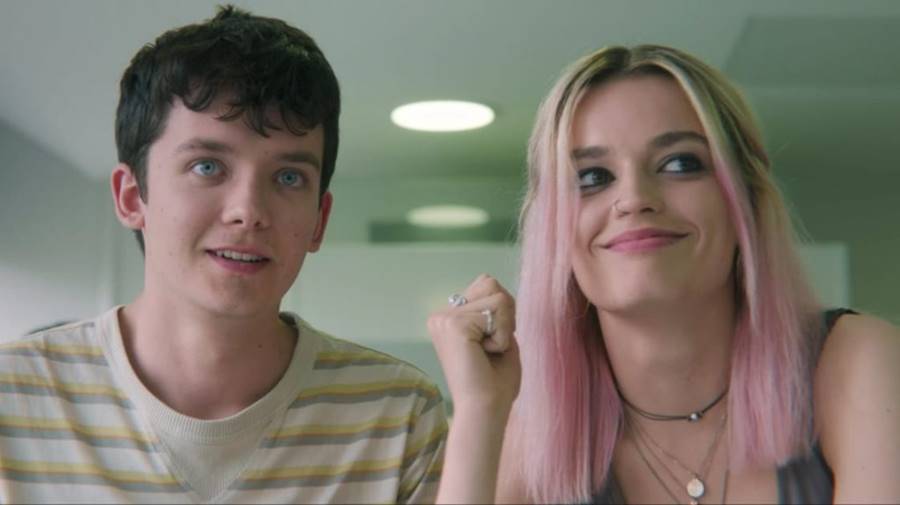 Foto colorida de um jovem com camiseta listrada e uma jovem com cabelo rosa um ao lado do outro sorrindo olhando para o lado- Metrópoles