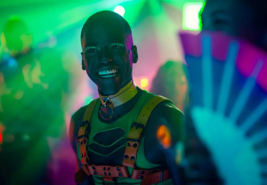 Foto colorida de um jovem negro sorrindo com roupas e maquiagem fluorescentes em um fundo com luzes coloridas - Metrópoles