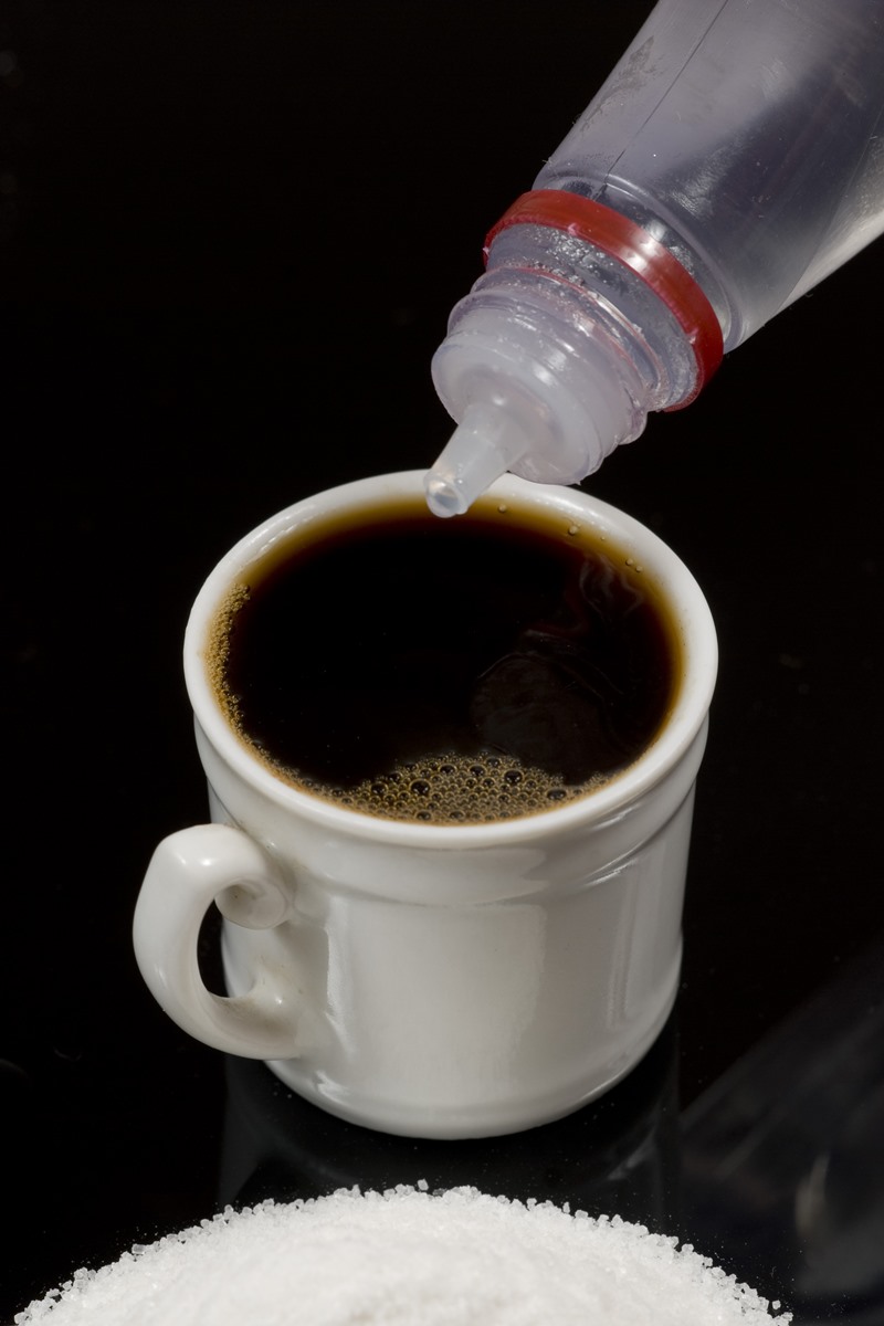 imagem colorida. Uma xícara de café está sobre um fundo escuro e uma mão aplica o adoçante - Metrópoles