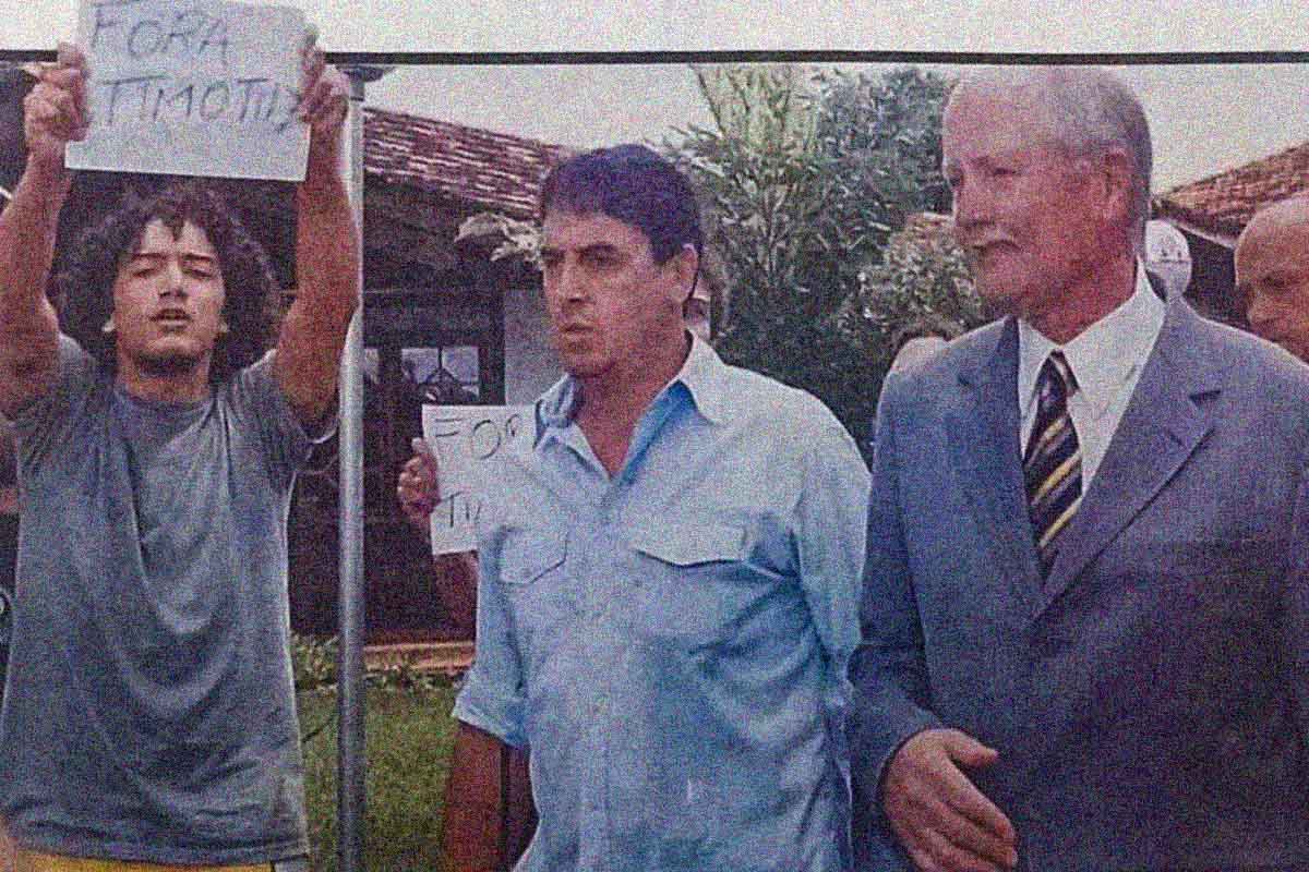 Timothy de terno e estudantes ao lado seguram cartaz exigindo saída do reitor 