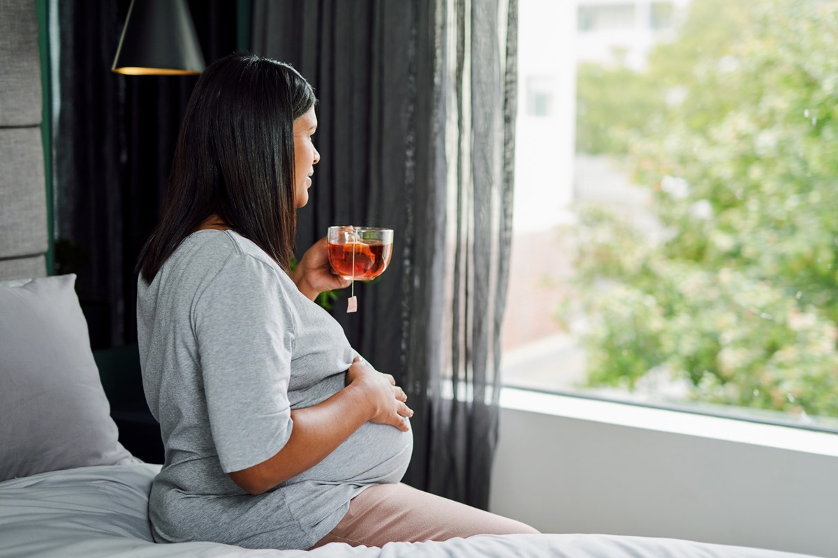 Foto colorida de mulher grávida sentada em uma cama e olhando a vista de uma janela. Ela segura uma xícara de chá - Metrópoles