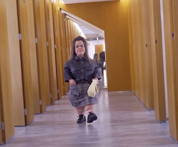 Em provador de roupas, mulher com nanismo desfila usando camisa de couro, saia e sapato preto - Metrópoles