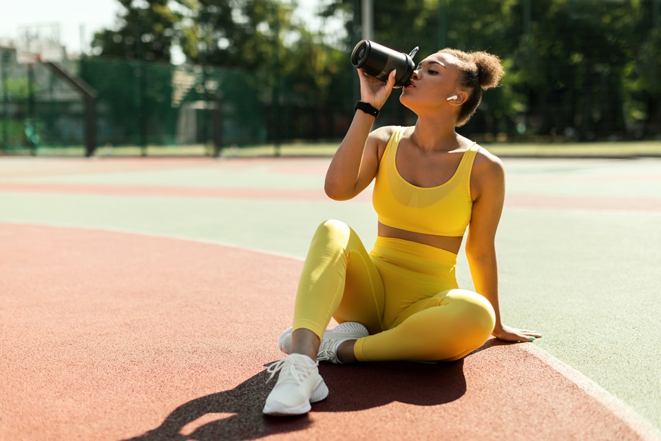 Mulher jovem sentada em quadra de esportes bebendo shake em garrafinha - Metrópoles