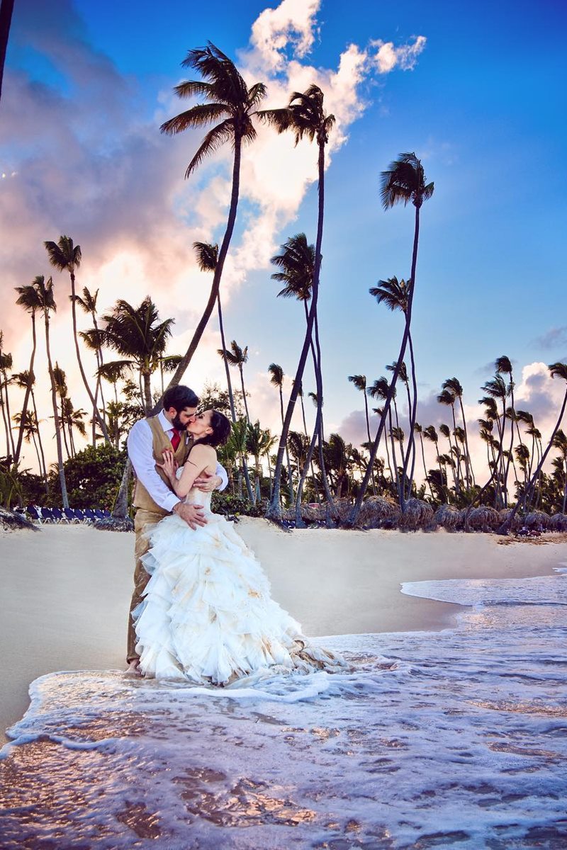 Foto colorida de homem e mulher, com roupas de noivos, se beijando enquanto a água do mar chega próximo a eles - Metrópoles