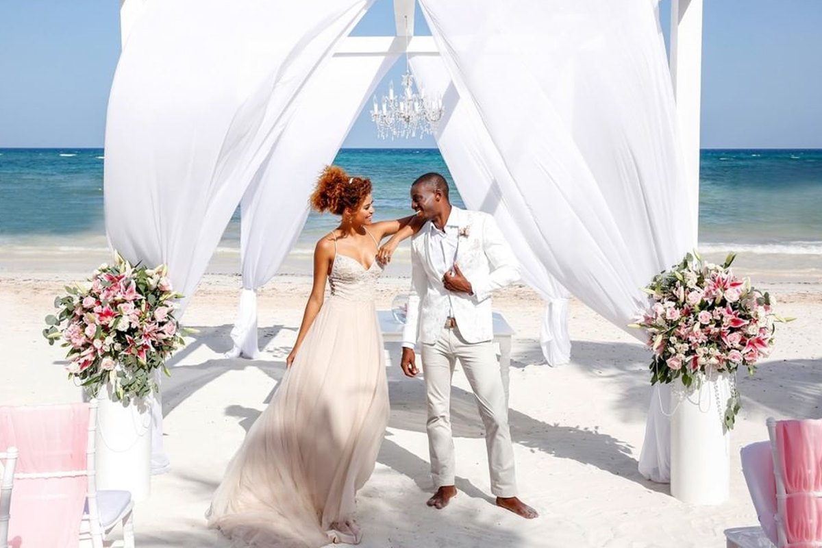 Foto colorida de mulher e homem, com roupa de noivos, em uma orla de praia com decoração de casamento - Metrópoles