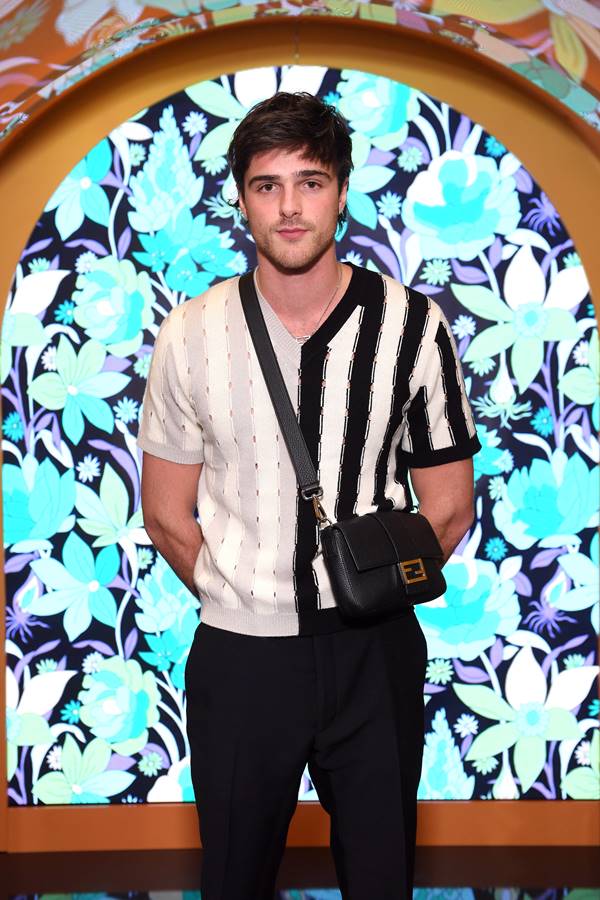 Em fundo floral, o ator Jacob Elordi usa blusa listrada em preto e branco, com bolsa preta de alça transpassada - Metrópoles