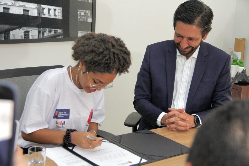 Imagem colorida mostra Isabela dos Santos, adolescente negra, de cabelos pretos e camiseta branca, assinando documento em uma mesa ao lado do prefeito Ricardo Nunes - Metrópoles