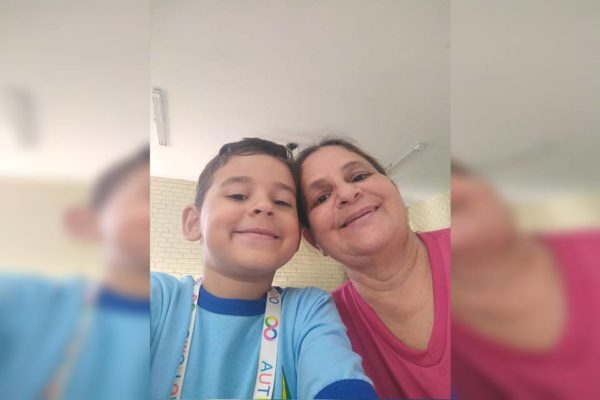 Foto colorida de criança com blusa azul e mulher branca com roupa rosa fazendo selfie e sorrindo 