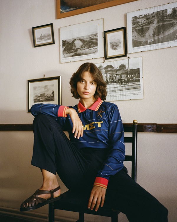 Mulher sentada em uma cadeira, com uma perna esticada e a outra apoiada na cadeira. Ela veste uma camisa de time azul e calça preta - Metrópoles