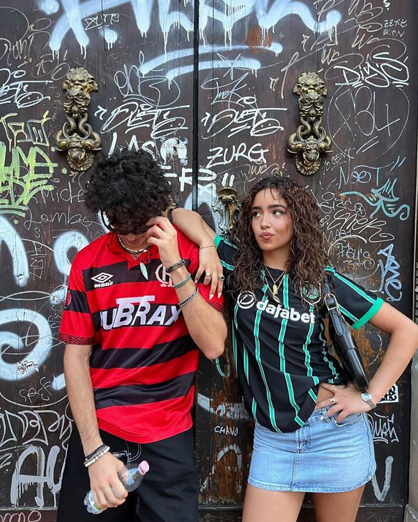 Dois adolescentes em frente a uma parede pichada, o homem vestindo uma camisa do Flamengo e a moça de outro time - Metrópoles