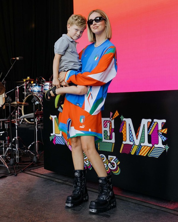 Influencer italiana Chiara Ferragni, loira e alta, veste camisa e shorts da seleção italiana, botas pretas e óculos escuros. Ela carrega seu filho nos braços - Metrópoles