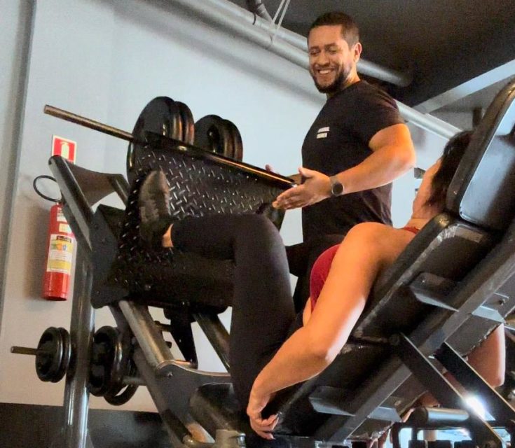 Homem sorrindo ao lado de um aparelho de academia, com uma mulher praticando leg press