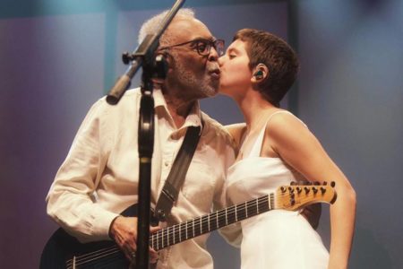 Foto colorida do cantor Gilberto Gil dando um beijo na neta, e também cantora, Flor Gil. Os dois vestem roupas brancas, e ele segura um violão nas mãos - Metrópoles