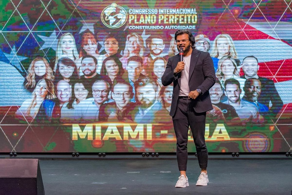 Foto colorida de homem em um palco, com telão ao fundo. Ele segura um microfone - Metrópoles