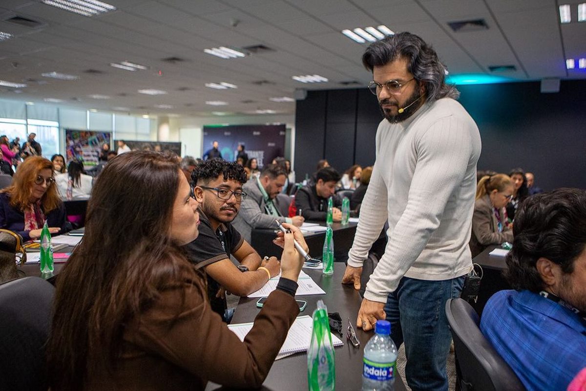 Foto colorida de homem em pé falando com duas mulher que está sentada ao redor de outras pessoas - Metrópoles