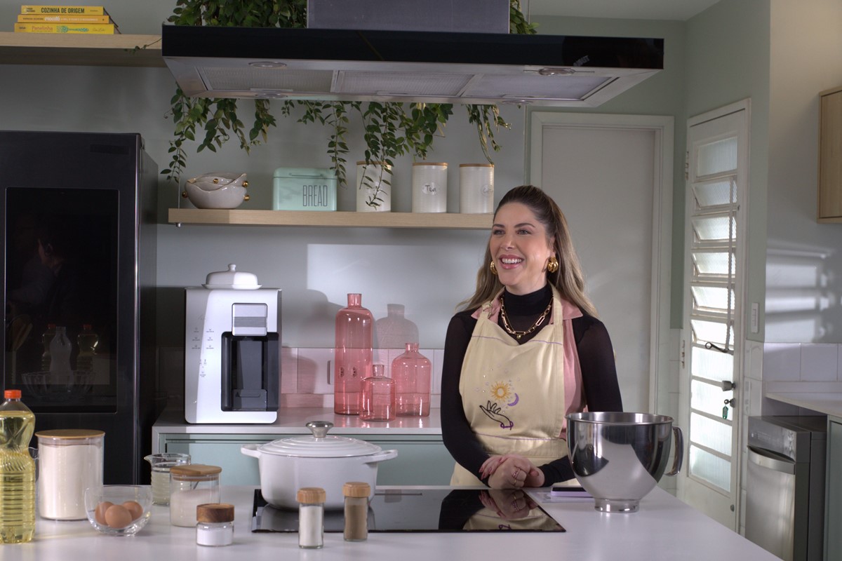 Foto colorida de mulher loira em cozinha com tons pastel. Ela usa um avental - Metrópoles