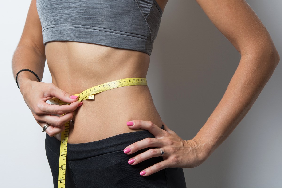 Foto colorida de mulher com fita métrica amarela na barriga e mão na cintura para representar perda de peso - Metrópoles