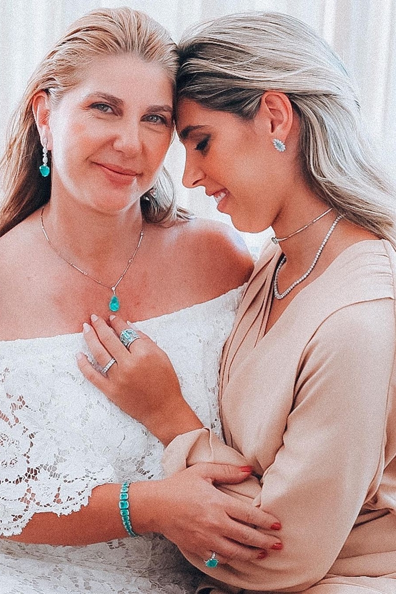 Foto colorida de mulher madura, loira e com joias na cor azul turquesa. Ao lado, uma mulher jovem e loira apoia a cabeça dela - Metrópoles