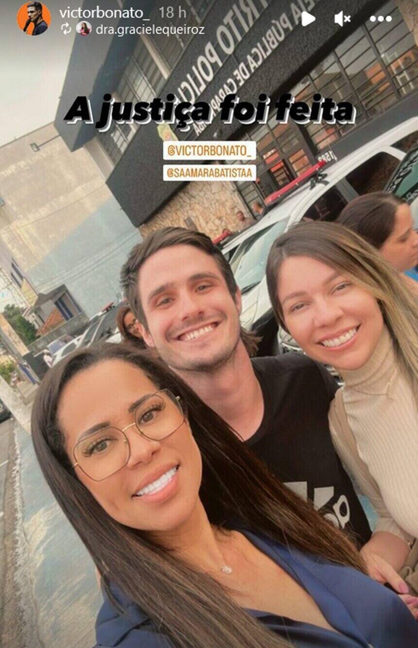 Em foto colorida homem branco sorri tendo ao seu lado esquerdo uma jovem loira e no direito uma morena, ambas tambpem sorrindo