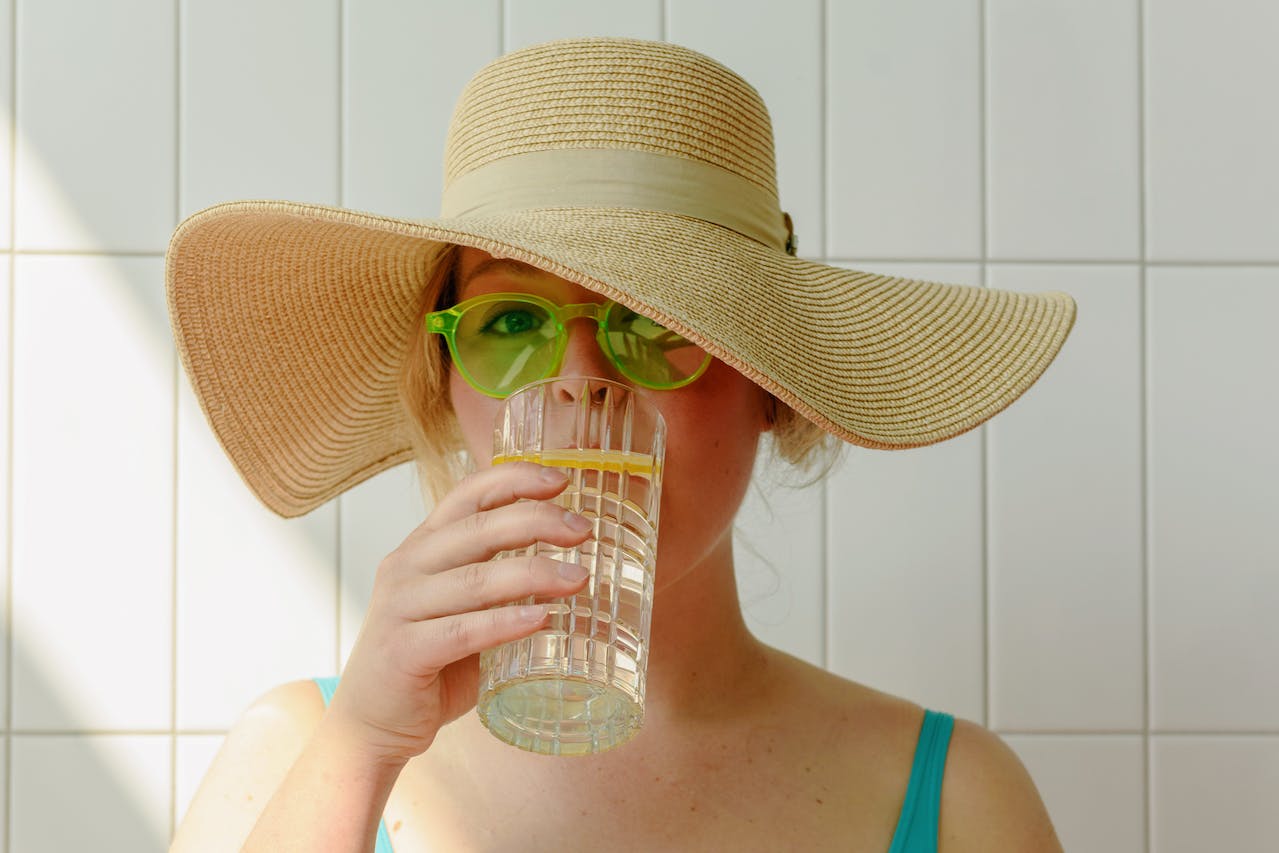 Jovem mulher de chapéu e óculos bebendo copo de água - Metrópoles