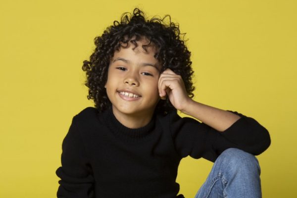 Foto de um menino negro, com o cabelo preto e grande, usando um moletom preto. O fundo é amarelo - Metrópoles