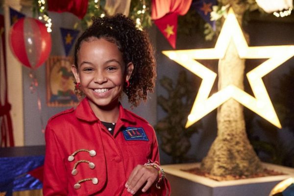 Foto de uma menina negra, com cabelo preto, sorrindo. Usa look vermelho. Ao fundo, uma estrela brilhante - Metrópoles