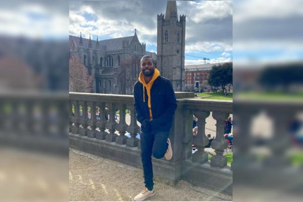 Foto colorida de homem negro com cascaco em um ponte com traços góticos de costas para a torre de um castelo