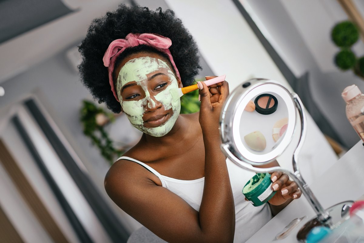 Foto colorida de mulher negra passando um creme verde no rosto enquanto olha para o espelho - Metrópoles