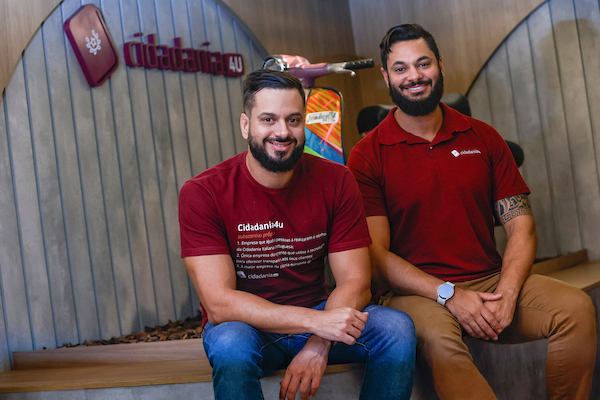 Fotografia colorida mostrando dois homens de camisa vermelha sentados em palco-Metrópoles