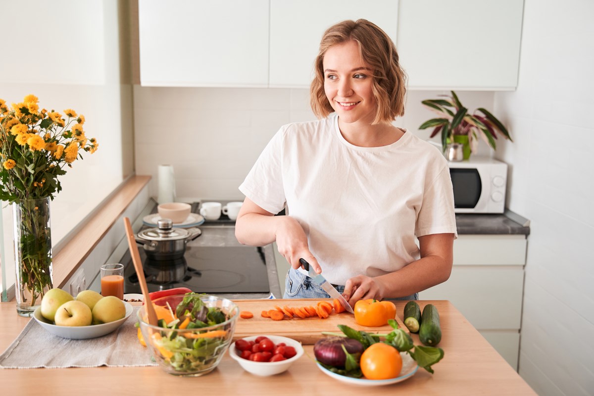 Foto colorida de mulher loira cortando cenoura. Ela está atrás de uma bancada com legumes - Metrópoles