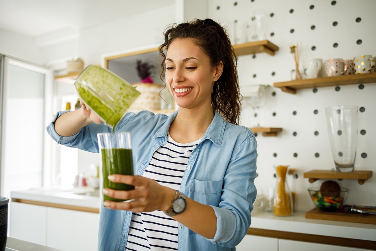 Mulher branca, de cabelos pretos prepara smoothie verde em casa - Metrópoles