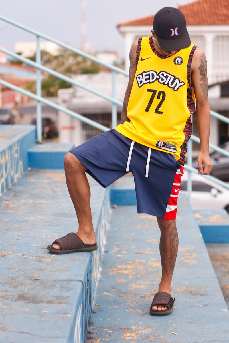 Homem em escadaria vestindo camiseta regata de basquete amarela, bermuda azul escuro com detalhes em branco e vermelho e sandálias pretas.