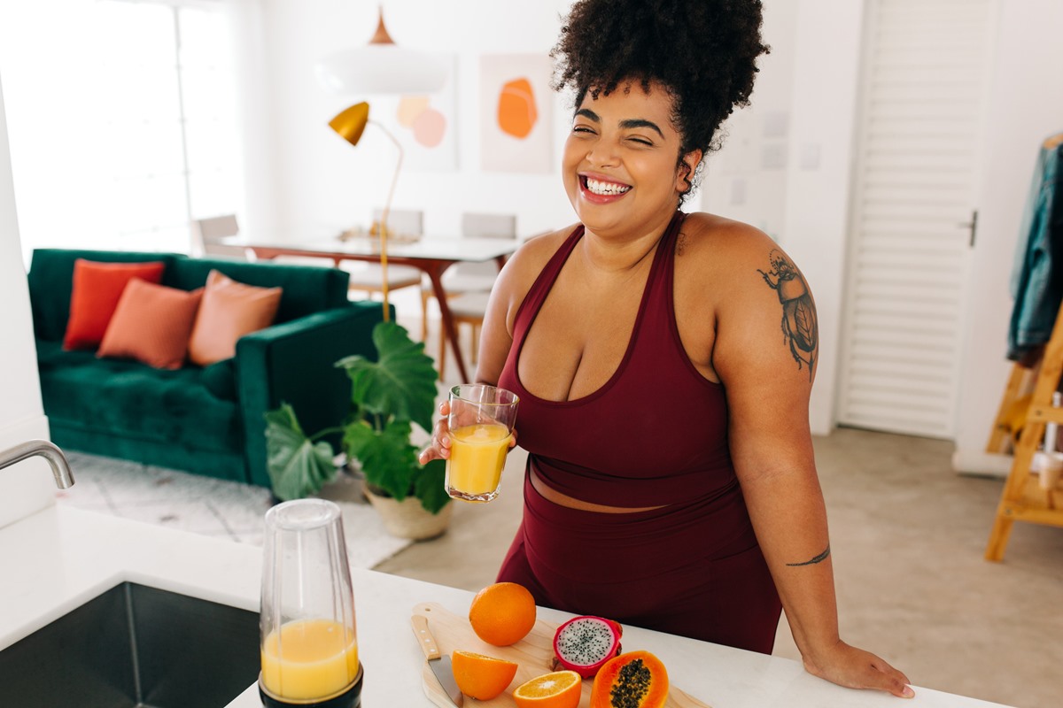 Foto colorida de mulher com roupa de malhar na cor vinho. Ela segura um copo de suco e está próximo de uma bancada com frutas - Metrópoles