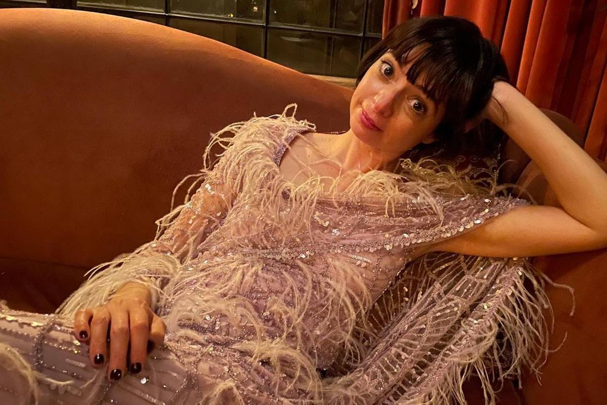 Kate Micucci deitada em sofá, com a mão na cabeça e usando vestido de plumas - Metrópoles