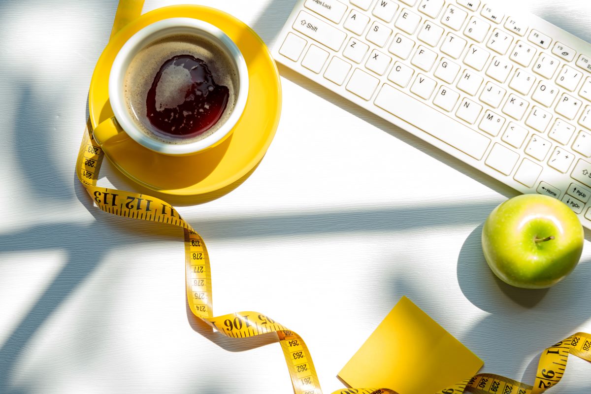 Foto de xícara amarela com café dentro. Ao lado tem uma fita métrica, um teclado de computador branco e uma maçã verde - Metrópoles