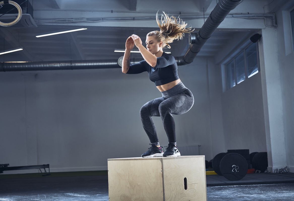 Mulher branca, adulta, loira está pulando de cima de uma caixa. Para fazer o exercício ela está usando uma roupa de academia toda preta - Metrópoles