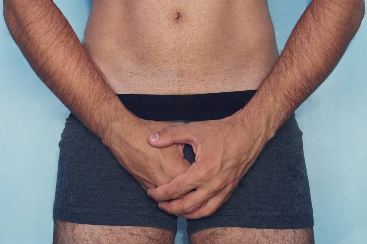 Foto colorida de um homem da cintura para baixo de cueca e cobrindo o pênis em um fundo azul - Metrópoles