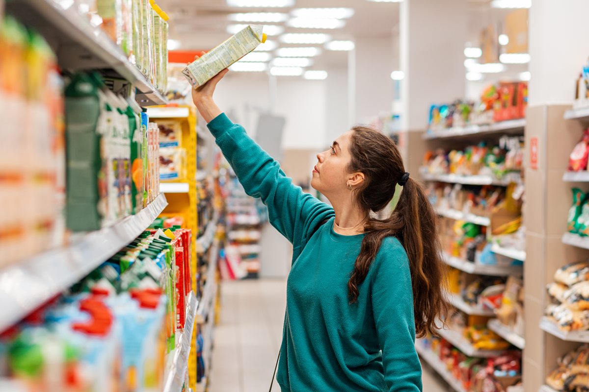 Foto colorida de mulher retirando uma caixa de suco de uma prateleira. Ela está em um supermercado - Metrópoles