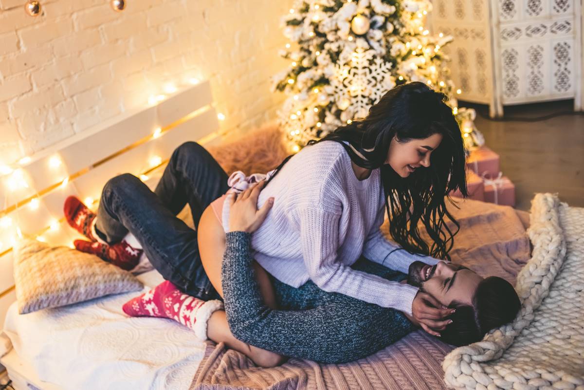 Foto colorida de um casal deitado em cima de uma cama com uma árvore de Natal do lado - Metrópoles