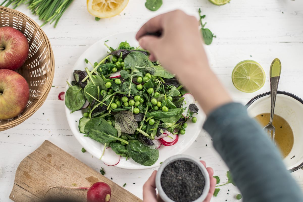 Foto colorida de prato com vegetais e mão feminina adicionando ervilhas. Do lado do prato, há duas maçãs e uma tigela com especiarias - Metrópoles