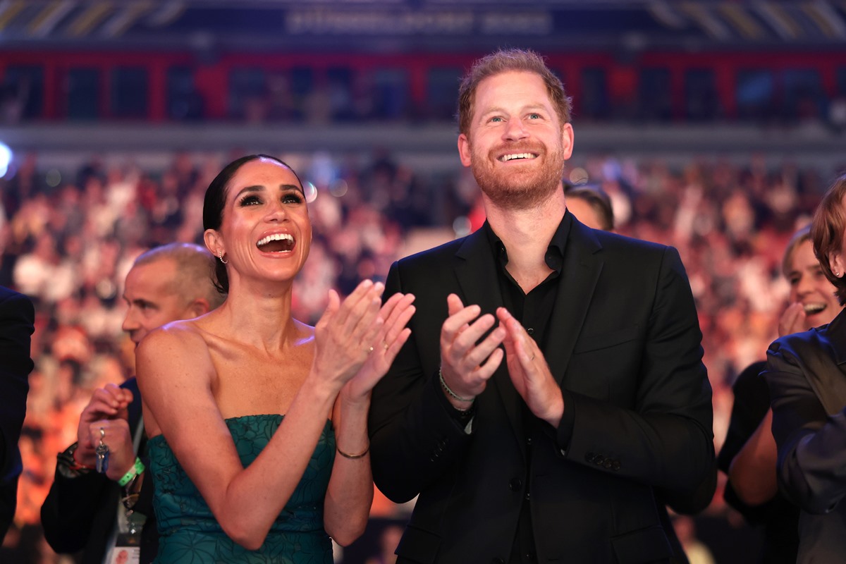 Harry e Meghan estão batendo palmas e sorrindo durante o Jogo Invictus. Ela é uma mulher negra e está com cabelos amarrados em coque e trajando um vestido verde. Harry é um homem branco, ruivo, que está usando um terno todo preto - Metrópoles