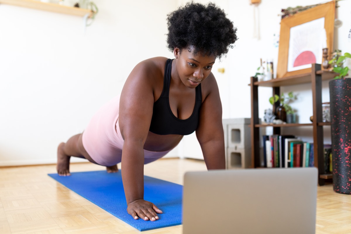 Foto colorida de mulher fazendo flexão em tapetinho enquanto olha para o notebook - Metrópoles