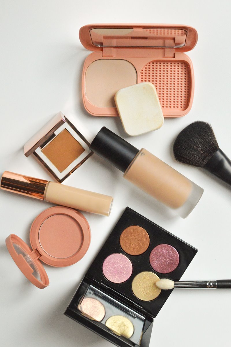 Produtos de maquiagem como blush, sombra, base, corretivo e pincel espalhados em um fundo branco