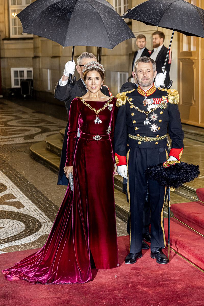 Foto colorida de mulher, com tiara, medalhas e vestido vermelho, ao lado de homem com trajes militares. Eles estão cobertos por guarda-chuva - Metrópoles 