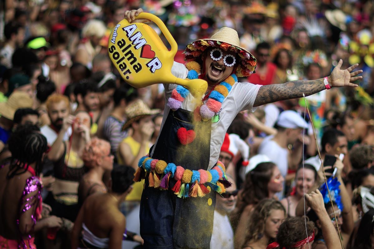 Homem está segurando um regador de planta e usando adereços coloridos durante o carnaval do Rio de Janeiro. Ele está usando óculos escuro, chapéu de palha e sorrindo pra foto - Metrópoles