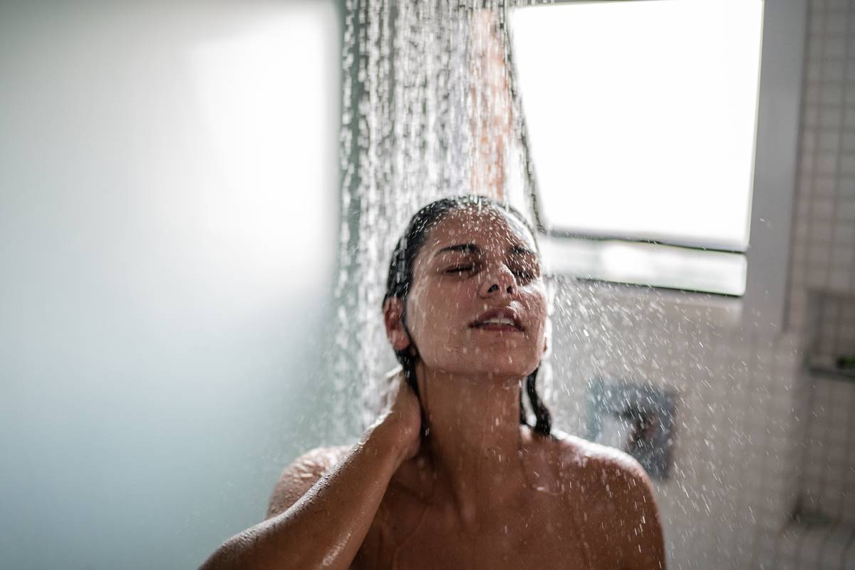 Foto colorida de uma mulher no chuveiro tomando banho de olhos fechados - Metrópoles