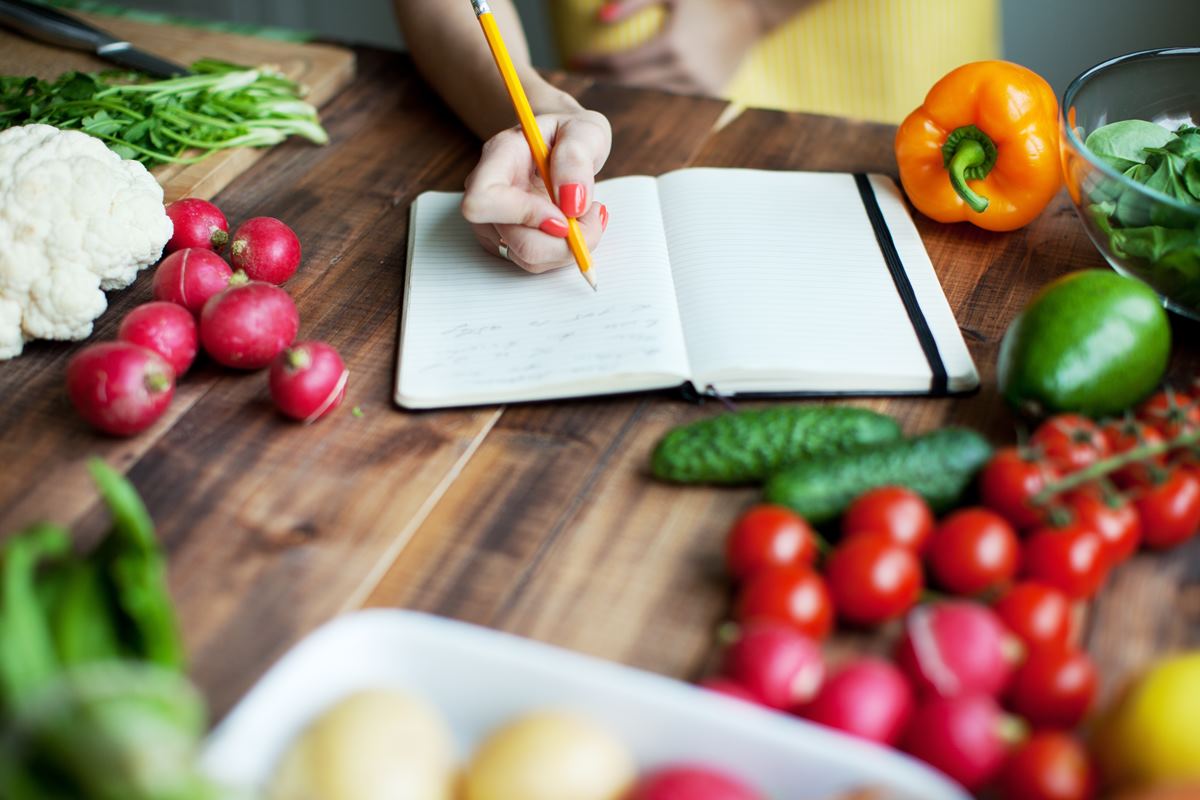 Imagem colorida mostra uma mesa marrom com vários verduras em diferentes cores. No meio, tem a mão de uma mulher anotando algo em uma carderno - Metrópoles