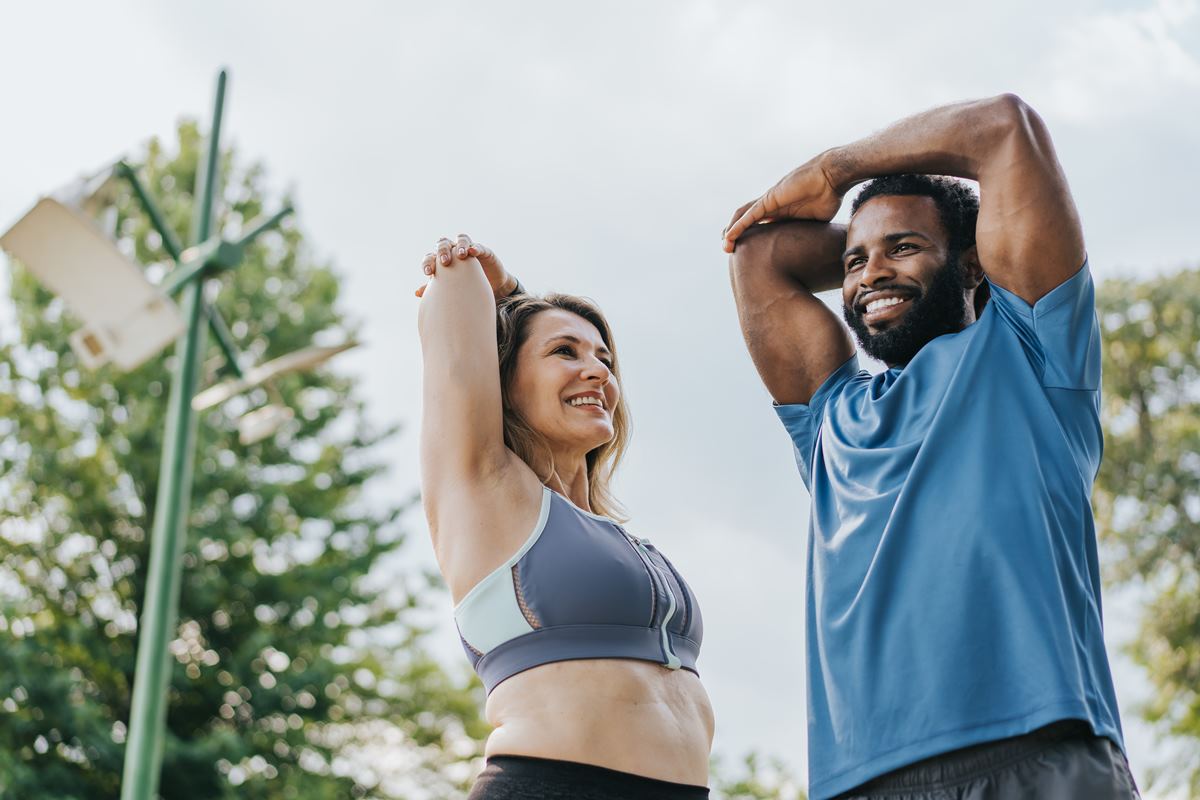 Mulher branca e homem alto, negro estão se alongando para fazer exercícios físicos, que ajudam a ativar o metabolismo - Metrópoles