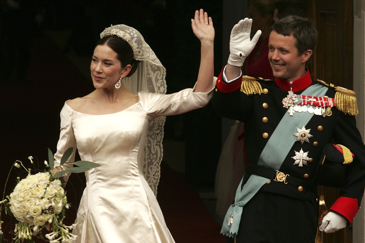 Foto colorida de mulher, com vestido de noiva e segurando buquê, ao lado de homem com trajes militares. Eles estão acenando - Metrópoles