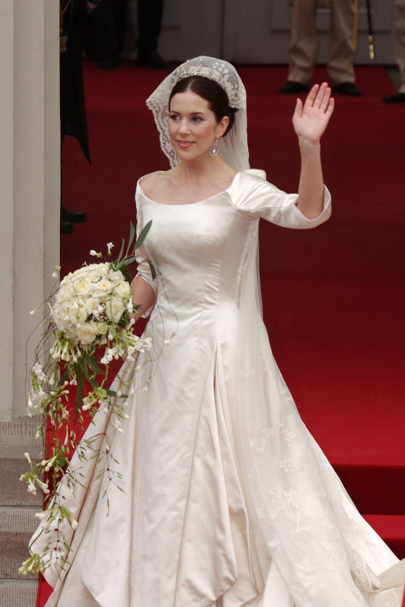 Foto colorida de mulher com vestido de noiva e segurando buquê. Ela está acenando - Metrópoles